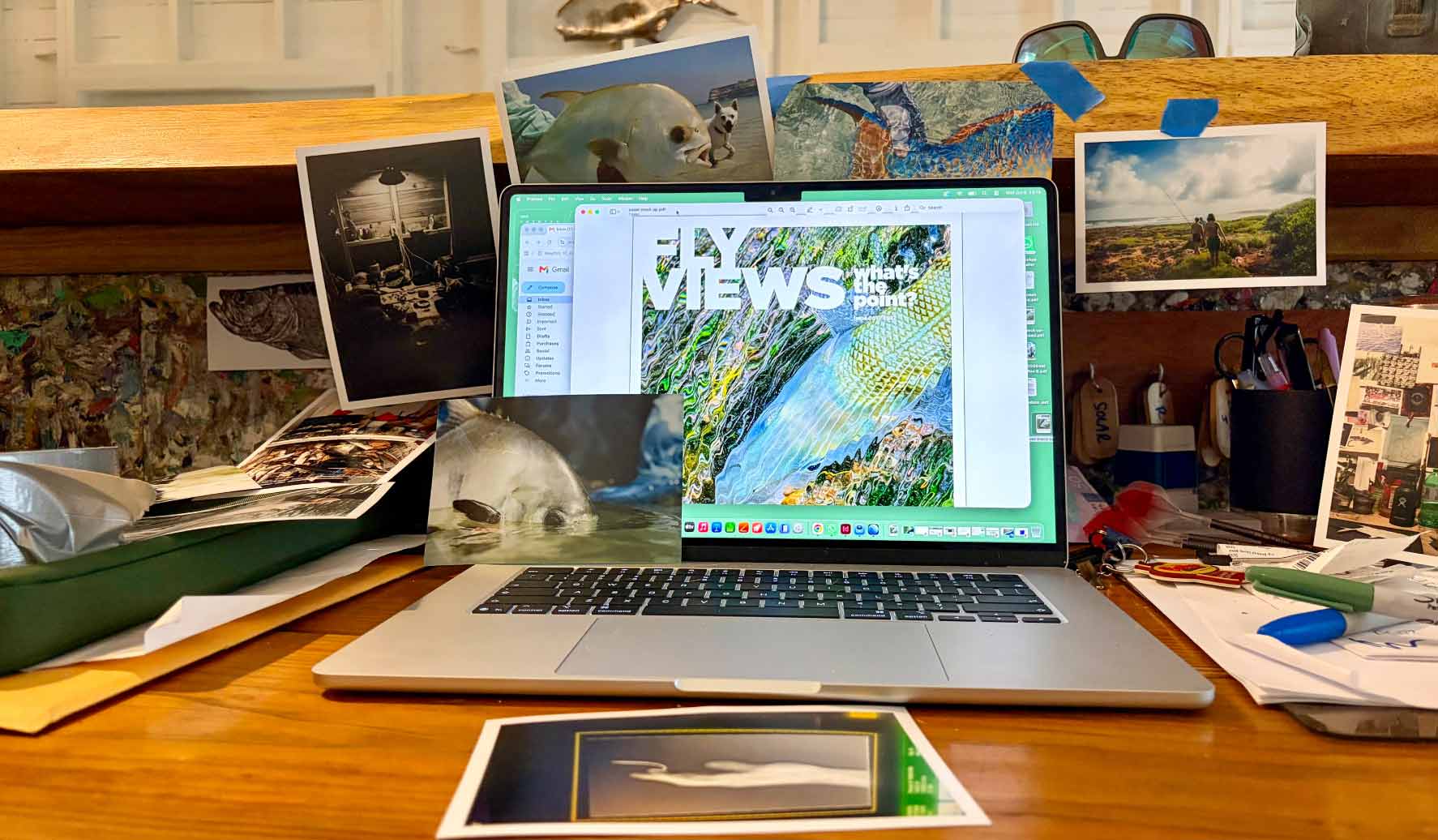 A creative desk with a laptop, artwork, photographs, and fly-fishing material.